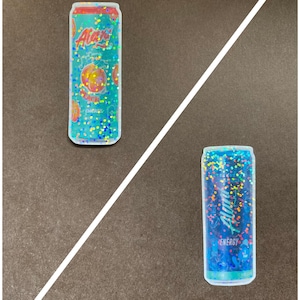 May include: Two aluminum cans with holographic glitter designs. The can on the left is labeled "Alani Nu" and "Juicy Peach" with a peach graphic. The can on the right is labeled "Alani Nu" and "Breezyberry" with a blue and purple berry graphic.