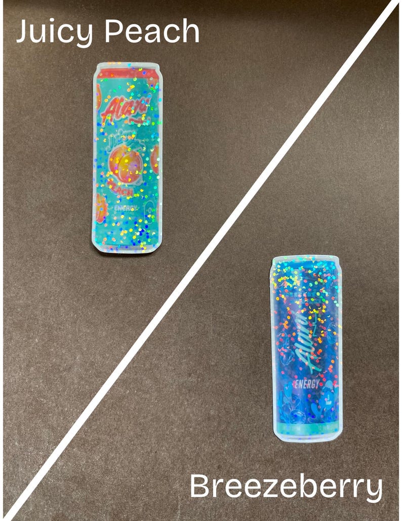 May include: Two aluminum cans with holographic glitter designs. The can on the left is labeled "Alani Nu" and "Juicy Peach" with a peach graphic. The can on the right is labeled "Alani Nu" and "Breezyberry" with a blue and purple berry graphic.