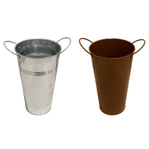 Może przedstawiać: Dwa metalowe wiaderka na kwiaty, jedno srebrne, a drugie brązowe. Oba wiaderka mają uchwyt i są cylindryczne.