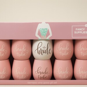 A pastel pink bridal gift box displayed in a studio setting, containing eight stemless wine glasses. Seven are soft pink with the words “bride tribe” in elegant white cursive, and one is white with the word “bride” in gold script. The packaging features a turquoise floral-patterned dress illustration and “Fancy Supplies” branding. Soft professional lighting highlights the smooth surfaces and pastel tones.