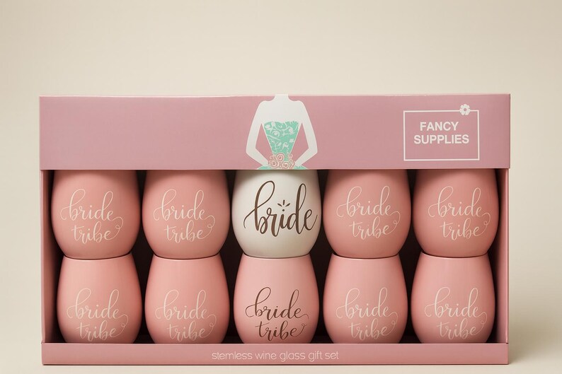 A pastel pink bridal gift box displayed in a studio setting, containing eight stemless wine glasses. Seven are soft pink with the words “bride tribe” in elegant white cursive, and one is white with the word “bride” in gold script. The packaging features a turquoise floral-patterned dress illustration and “Fancy Supplies” branding. Soft professional lighting highlights the smooth surfaces and pastel tones.