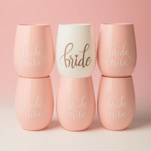 A set of six pastel-colored stemless wine glasses arranged in two rows. Five glasses are light pink with the words “bride tribe” in white cursive, and the center glass is white with the word “bride” in elegant brown script featuring a small diamond design. The setup is in a studio with soft pink background and polished lighting.