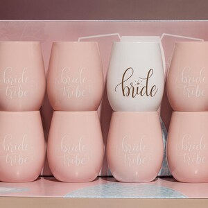 A studio photograph of six pastel-colored stemless wine glasses arranged in two rows. Five are light pink with the words “bride tribe” in elegant white cursive, and one central glass is white with the word “bride” in gold script. Shot from a side angle, the image highlights depth, gloss, and soft reflections against a bridal-themed pink backdrop.