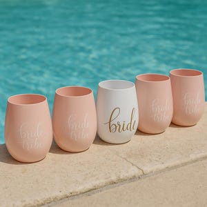 A set of five blush pink stemless wine glasses with “bride tribe” in white cursive and one central white glass with “bride” in gold script, arranged neatly on the beige stone edge of a swimming pool. The turquoise water sparkles in the background, creating a bright, summery lifestyle scene without people.