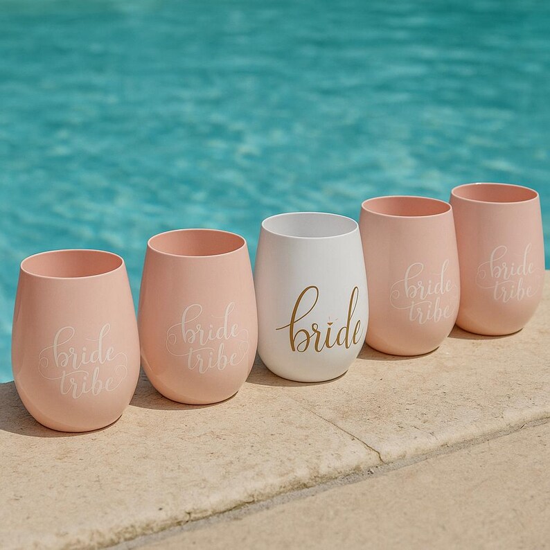 A set of five blush pink stemless wine glasses with “bride tribe” in white cursive and one central white glass with “bride” in gold script, arranged neatly on the beige stone edge of a swimming pool. The turquoise water sparkles in the background, creating a bright, summery lifestyle scene without people.