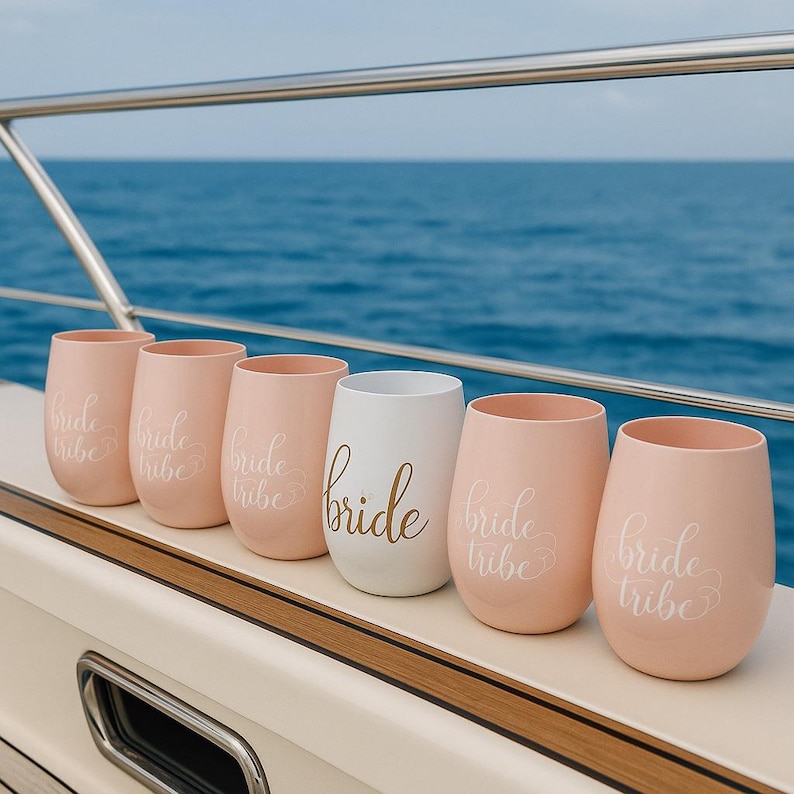 A row of six stemless wine glasses placed neatly on the deck of a yacht. Five are blush pink with “bride tribe” in white cursive, and one central glass is white with “bride” in gold script. The scene shows a stainless-steel railing and the open ocean in the background, with soft daylight highlighting the nautical lifestyle setting. No people present.