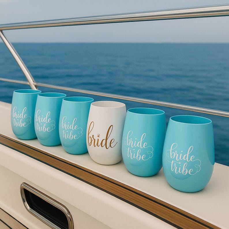 A row of six stemless wine glasses placed neatly on the deck of a yacht. Five are blush blue with “bride tribe” in white cursive, and one central glass is white with “bride” in gold script. The scene shows a stainless-steel railing and the open ocean in the background, with soft daylight highlighting the nautical lifestyle setting. No people present.