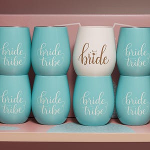 A studio photograph of six pastel-colored stemless wine glasses arranged in two rows. Five are light blue with the words “bride tribe” in elegant white cursive, and one central glass is white with the word “bride” in gold script. Shot from a side angle, the image highlights depth, gloss, and soft reflections against a bridal-themed pink backdrop.