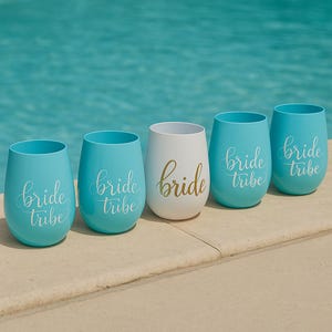 A set of five blush blue stemless wine glasses with “bride tribe” in white cursive and one central white glass with “bride” in gold script, arranged neatly on the beige stone edge of a swimming pool. The turquoise water sparkles in the background, creating a bright, summery lifestyle scene without people.