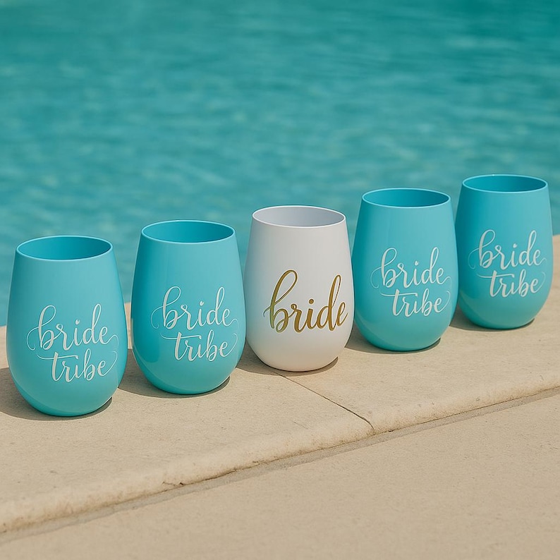 A set of five blush blue stemless wine glasses with “bride tribe” in white cursive and one central white glass with “bride” in gold script, arranged neatly on the beige stone edge of a swimming pool. The turquoise water sparkles in the background, creating a bright, summery lifestyle scene without people.