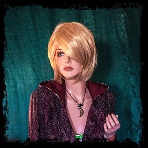 May include: A mannequin head with a short, blonde wig. The mannequin is wearing a burgundy paisley print jacket with a red hood. A necklace with a crescent moon pendant and beads is visible. The background is a dark teal color.
