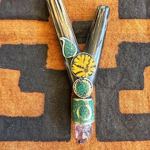 May include: A Y-shaped, handcrafted smoking pipe with a dark, striped finish. It features decorative elements including green and yellow circular inlays, and a raw amethyst crystal at the base. The pipe rests on a patterned orange and black fabric.