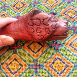 May include: A hand-carved wooden sculpture of a jaguar head. The sculpture is a rich brown color and features detailed carvings of the jaguar's facial features and spots. The jaguar is held over a colorful, patterned textile.