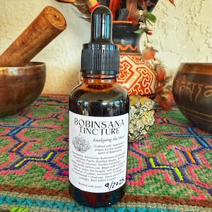 以下が含まれることがあります： 黒いスポイトが付いた、ボビンサナチンクチャーの茶色のガラス瓶。ラベルには「Awakening the Heart」と書かれています。ボトルはカラフルな模様の表面にあり、背景には他の装飾品があります。有効期限は9/2025です。