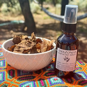 May include: A brown glass spray bottle labelled "Amazonian Mapacho Spray" beside a white bowl filled with dried leaves. The bottle has a black cap and a clear spray nozzle. The items are on a colourful, patterned surface.