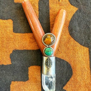May include: A wooden Y-shaped pipe with a clear quartz crystal at the base. The pipe features a silver band with a leaf design and two gemstone accents: a tiger's eye and a turquoise stone. The background is a black and orange patterned fabric.