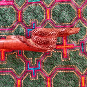 Puede incluir: Un mango de bastón de madera de color marrón rojizo tallado con la forma de una serpiente enroscada con texturas de escamas detalladas. La cabeza de la serpiente está situada en el extremo, con ojos tallados y un hocico liso.