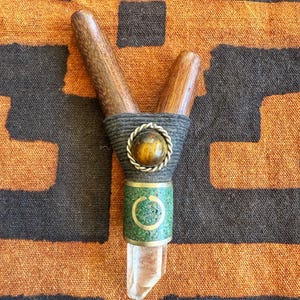 May include: A handcrafted Y-shaped tool featuring wooden handles, a wrapped center with a decorative stone, and a clear crystal point. The tool has a green and gold band near the crystal. The background is a patterned orange and black fabric.