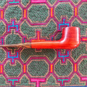 May include: A tobacco pipe featuring a smooth, reddish-brown wooden bowl and shank connected to a tapered, dark brown and tan patterned stem. The pipe is positioned horizontally, showcasing the contrast between the solid wood and the textured stem.