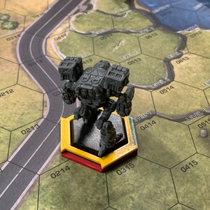May include: A gray miniature mech figure stands on a yellow and red hexagonal base, positioned on a game board with a road and terrain features. Another black figure is in the background. The board has numbered hexagonal cells.