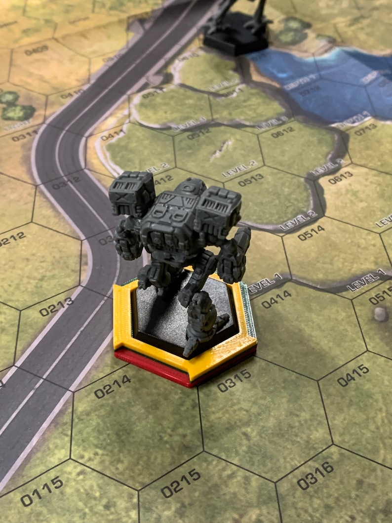 May include: A gray miniature mech figure stands on a yellow and red hexagonal base, positioned on a game board with a road and terrain features. Another black figure is in the background. The board has numbered hexagonal cells.