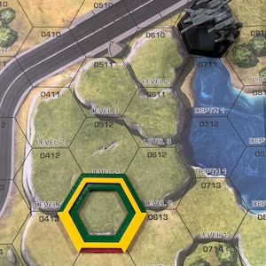 May include: A board game with a hexagonal grid, featuring terrain markers and a miniature gray vehicle. The game board has a road, water, and numbered hexes. A yellow, green, and red hexagonal marker is in the foreground.