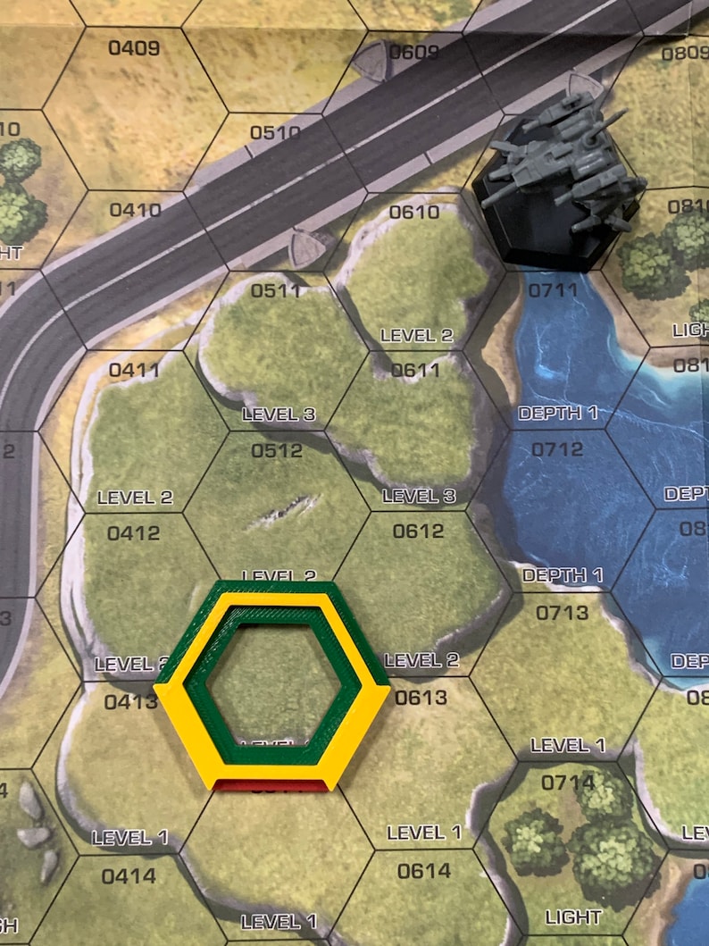 May include: A board game with a hexagonal grid, featuring terrain markers and a miniature gray vehicle. The game board has a road, water, and numbered hexes. A yellow, green, and red hexagonal marker is in the foreground.