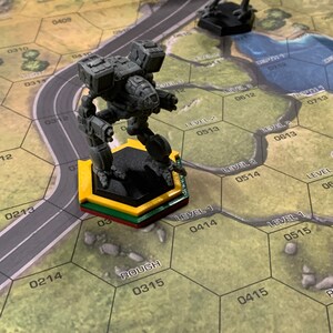 May include: A gray miniature robot figure stands on a yellow, green, and red hexagonal base on a game board. Another black robot figure is in the background. The game board has a road and hexagonal grid with numbers and the word "ROUGH".