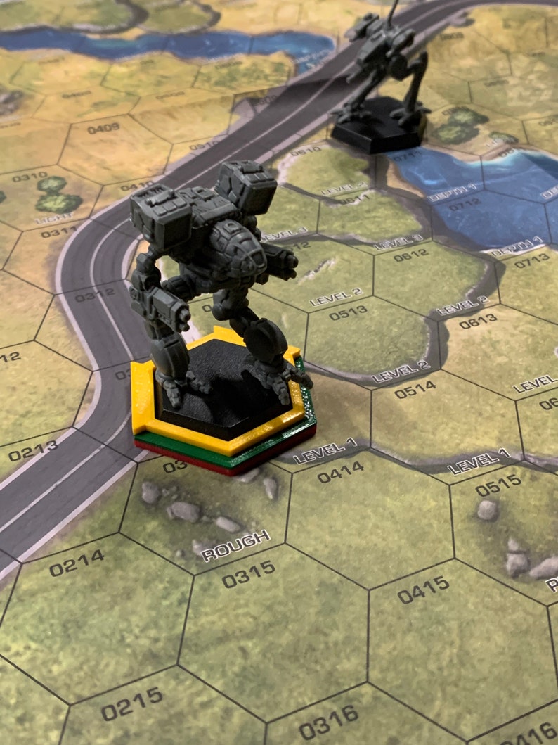 May include: A gray miniature robot figure stands on a yellow, green, and red hexagonal base on a game board. Another black robot figure is in the background. The game board has a road and hexagonal grid with numbers and the word "ROUGH".