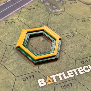 May include: A multi-layered, hexagonal game piece in yellow, green, and red, resting on a green and brown game board. The board features a road graphic and the word "ROUGH". The game piece is likely for a tabletop strategy game.