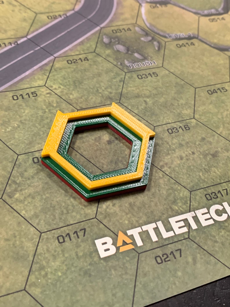 May include: A multi-layered, hexagonal game piece in yellow, green, and red, resting on a green and brown game board. The board features a road graphic and the word "ROUGH". The game piece is likely for a tabletop strategy game.