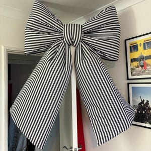May include: A large, decorative bow made of black and white striped fabric. The bow is mounted on a white wall and has long, flowing tails. The stripes are vertical and evenly spaced, creating a bold visual statement. The bow is a striking accent piece.