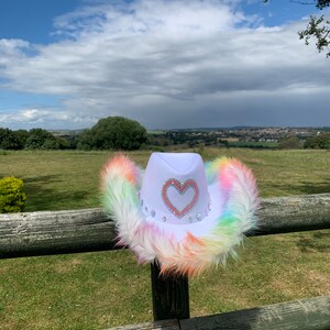 May include: A white cowboy hat with a rainbow colored faux fur trim and a pink heart with rhinestones on the front.