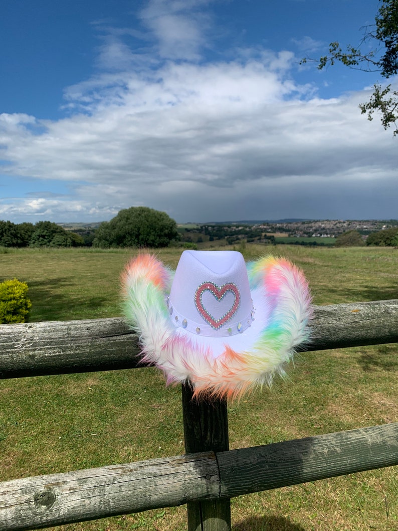 May include: A white cowboy hat with a rainbow colored faux fur trim and a pink heart with rhinestones on the front.