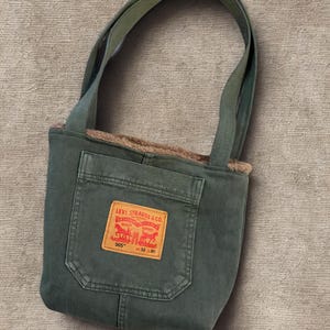 Op de afbeelding: Een groene tote bag met een zak en handvatten. De tas heeft een rood en geel Levi Strauss & Co. patch. De binnenkant is gevoerd met een bruin, fuzzy materiaal. De tas is gemaakt van een duurzame stof.