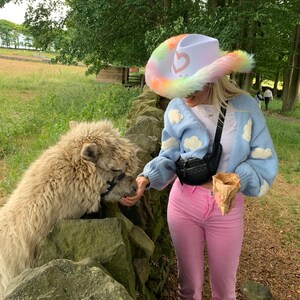 May include: A person wearing a white cowboy hat with a rainbow trim and a blue cardigan with white clouds printed on it, feeding a white alpaca. The person is wearing pink pants and holding a brown paper cone.