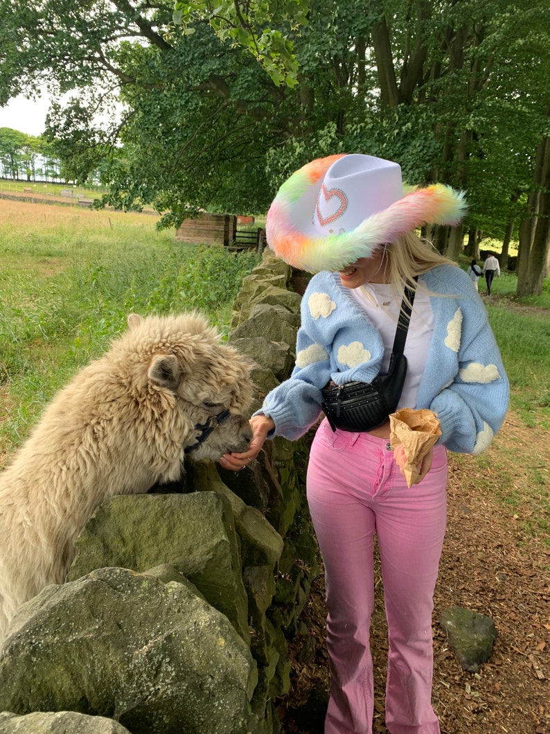 May include: A person wearing a white cowboy hat with a rainbow trim and a blue cardigan with white clouds printed on it, feeding a white alpaca. The person is wearing pink pants and holding a brown paper cone.
