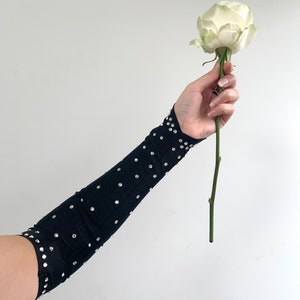 K&ouml;nnte beinhalten: Ein schwarzer Armschoner mit Strasssteinen, getragen am Arm einer Person, die eine einzelne wei&szlig;e Rose h&auml;lt.