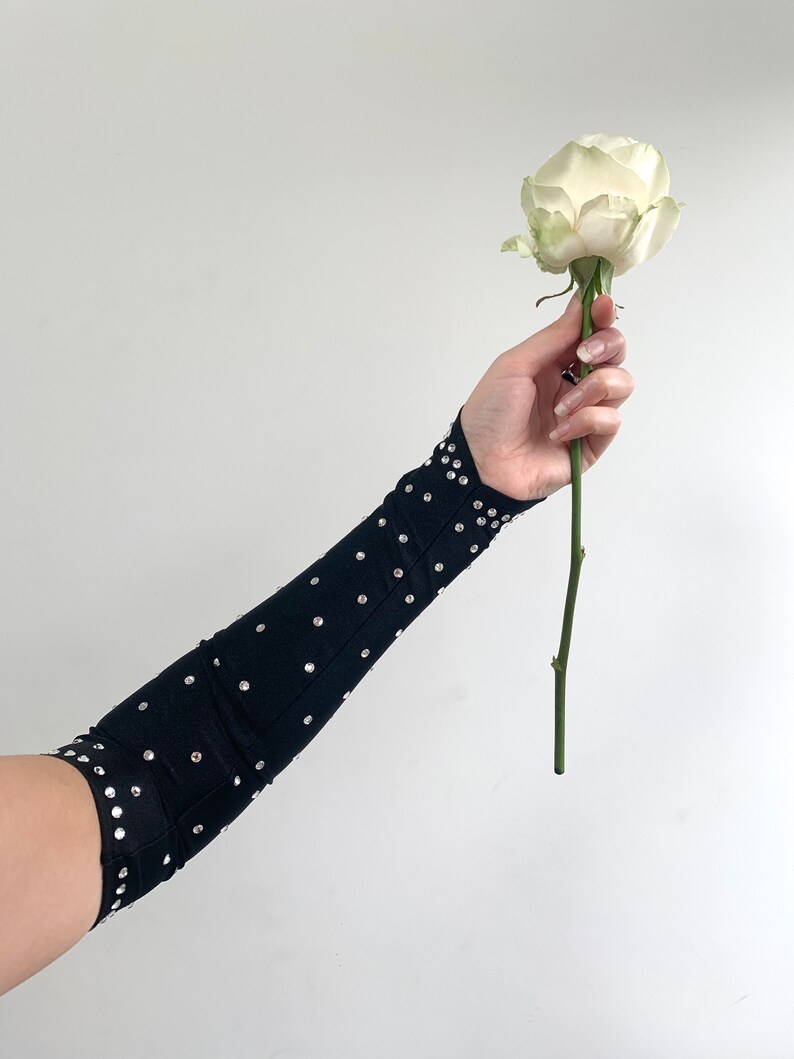 K&ouml;nnte beinhalten: Ein schwarzer Armschoner mit Strasssteinen, getragen am Arm einer Person, die eine einzelne wei&szlig;e Rose h&auml;lt.