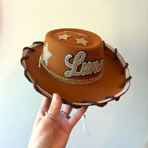 May include: A brown felt cowboy hat featuring the name Luna in cursive script, decorated with gold glitter stars on the crown, a gold braided trim, and a silver rhinestone border along the edge of the brim, accented with a dark brown twisted rope.