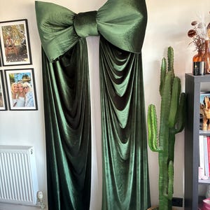 May include: A large, dark olive green velvet bow wall decoration featuring two long, draped fabric panels that extend downwards from the central knot, creating a soft, flowing appearance against the wall surface.