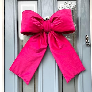 May include: A large, bright pink bow made of satin fabric. The bow is tied around a door handle and hangs down on either side of the door.