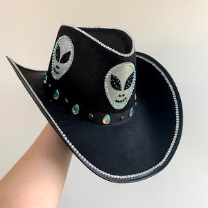 Op de afbeelding: Zwarte cowboyhoed met zilveren strass-afwerking en twee glitterende alien-gezichtapplicaties. De hoed heeft iriserende druppelvormige edelstenen en een zwarte band. Een uniek en stijlvol accessoire.