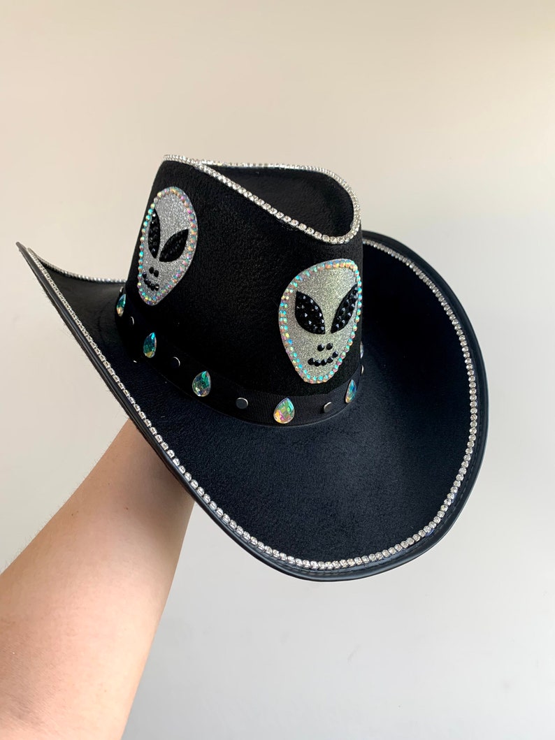 Op de afbeelding: Zwarte cowboyhoed met zilveren strass-afwerking en twee glitterende alien-gezichtapplicaties. De hoed heeft iriserende druppelvormige edelstenen en een zwarte band. Een uniek en stijlvol accessoire.