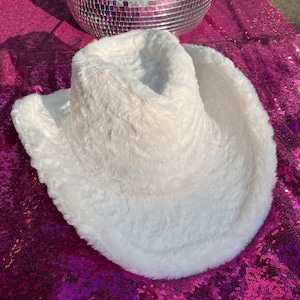 May include: A white faux fur cowboy hat with a wide brim. The hat is lying on a pink sequined surface.