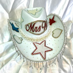 May include: White cowboy hat with decorative embellishments. Features a red star, heart, and bird accents. The hat is adorned with rhinestones, pearls, and the text "Mrs W". Silver fringe hangs from the brim.
