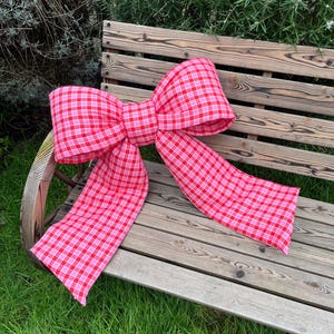 Puede incluir: Un gran lazo de gingham rosa y blanco sobre un banco de madera. El lazo está hecho de tela y tiene una forma grande y floja.