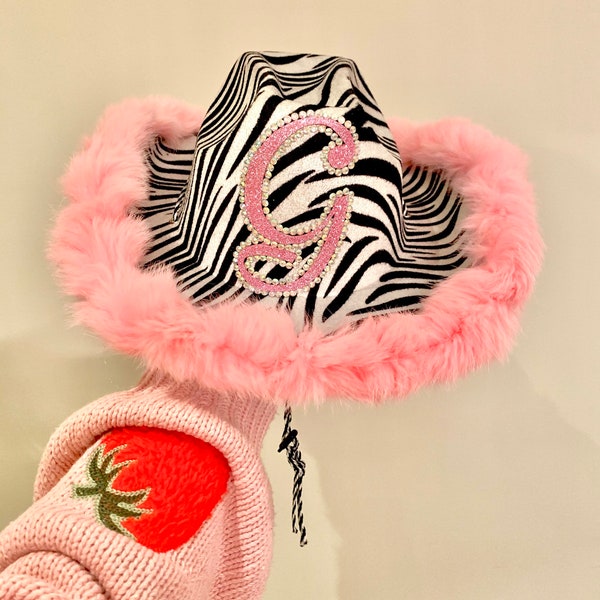 May include: A black and white zebra print cowboy hat with a pink faux fur trim. The hat has a pink glitter letter "G" on the front.