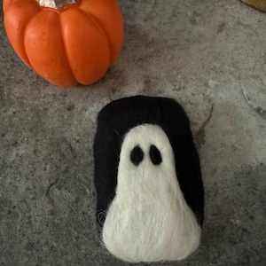 May include: A needle-felted ghost figure with a black background and a white ghost shape featuring two black oval eyes, positioned next to a small orange pumpkin with a silver stem. The ghost is crafted from soft, fuzzy fibres with a distinct texture.