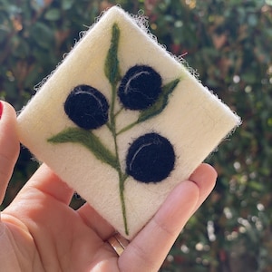 May include: A square, cream-coloured felt coaster with a branch of olives. The branch has green leaves and three black olives. The coaster is held in a hand against a blurred green background.